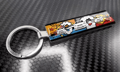 Llavero XL delgado estilo cómic con un diseño colorido de matrícula sobre una superficie con textura de carbono