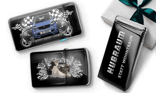Drei schwarze Geldbörsen mit Auto-Designs und karierten Flaggen, vor einem weißen Hintergrund. Eine zeigt ein Auto mit Rädern, eine andere ein Paar, und eine trägt den Text „Hubraum statt Wohntraum“.