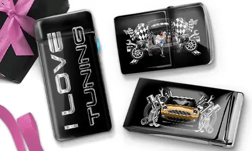 Drei glänzende Auto-Accessoires sind auf weißem Hintergrund zu sehen. Eines trägt die Aufschrift „I Love Tuning“, ein anderes zeigt Vintage-Rennmotive und das letzte ein blaues Sportauto mit Motor-Motiven. Eine rosa Schleife ist teilweise sichtbar.
