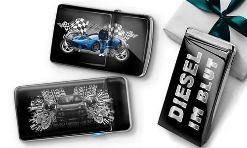 Two sleek black lighters featuring car themes, one with a blue car and checkered flags, another with a red car and gears. A third lighter reads 'DIESEL IM BLUT'. Gift-wrapped in the background.
