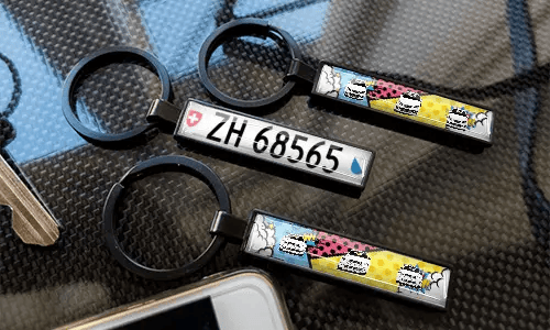 Schlüsselanhänger mit Nummernschild im Comic-Stil, präsentiert auf einer carbonstrukturierten Oberfläche mit farbenfrohen Auto-Designs